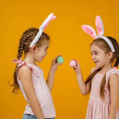 Zwei Kinder mit Hasenohren-Haarreifen stehen vor einem orangefarbenen Hintergrund und halten bunte Ostereier in den Händen.