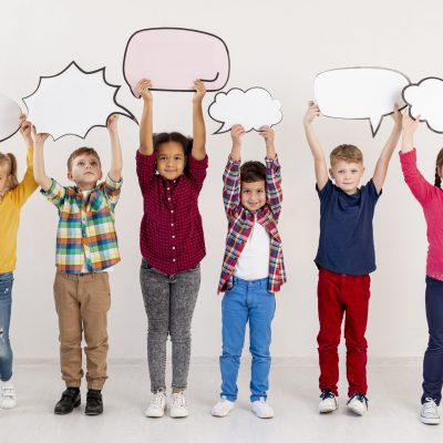 Sechs Kinder stehen nebeneinander und halten leere Sprechblasen aus Pappe über ihren Köpfen.