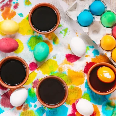 Mehrere bunt gefärbte Ostereier liegen auf Küchenpapier, daneben stehen kleine Schalen mit Eierfarbe und ein offener Eierkarton.