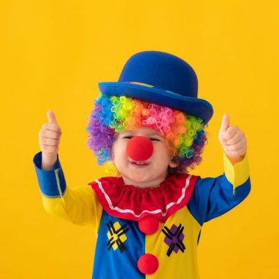 Funny kid clown showing thumbs up against yellow background. Happy child playing indoor. 1 April Fool's day concept