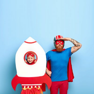 Super father wears mask, blue t shirt and red cloak, looks surprisingly into distance, plays with cute daughter, pretend being superheroes. Cheerful small kid keeps head in hole of toy rocket